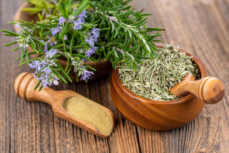 Fresh rosemary stems with small purple flowers next to a wooden bowl full of dried rosemary with a wooden scoop. An additional wooden scoop lays next to the fresh rosemary and wooden bowl. 