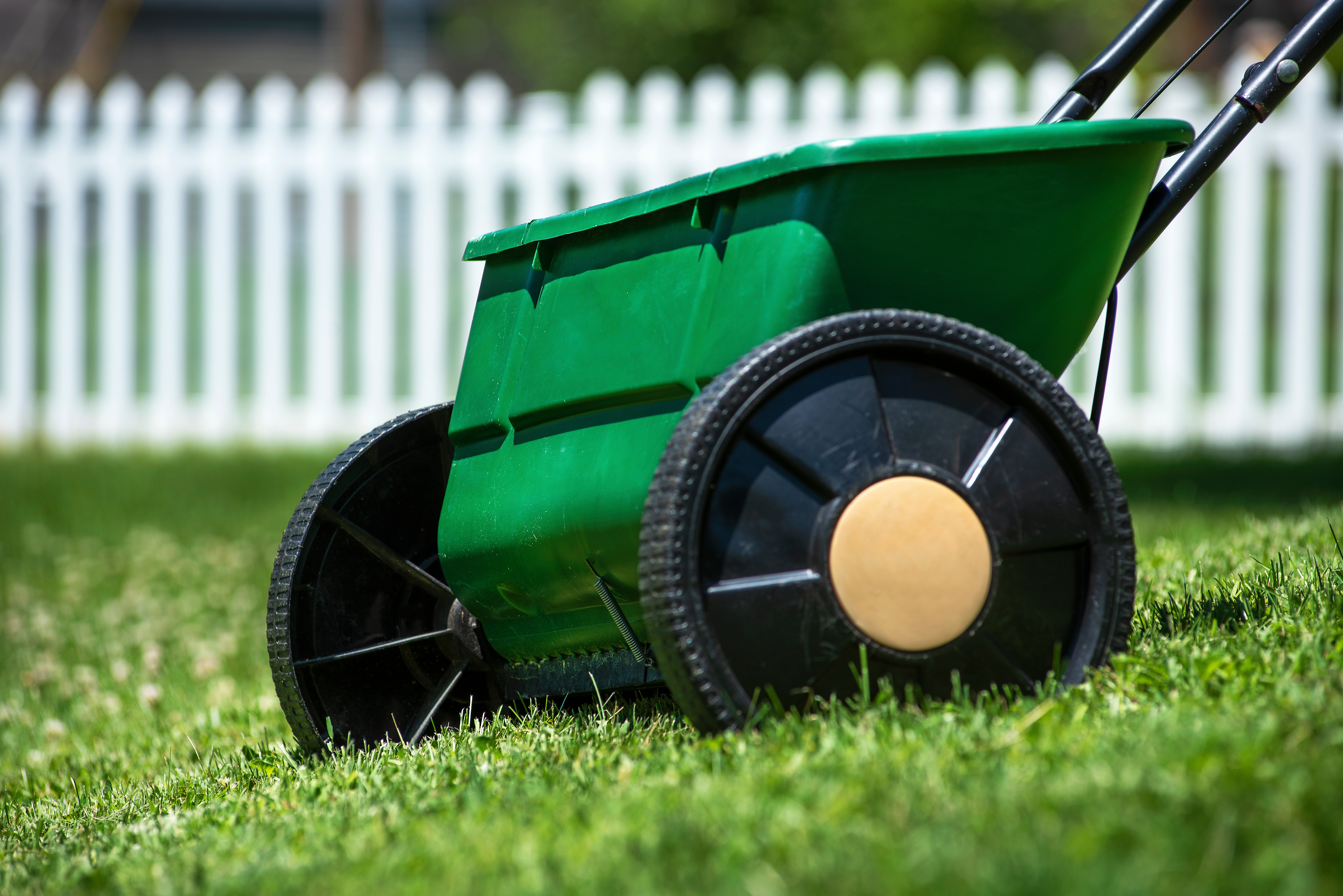 Drop seeder on green lawn near a white picket fence.