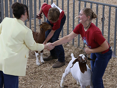 Woman in yellow blazer shakes hands with contestant holding a meat goat in a livestock arena