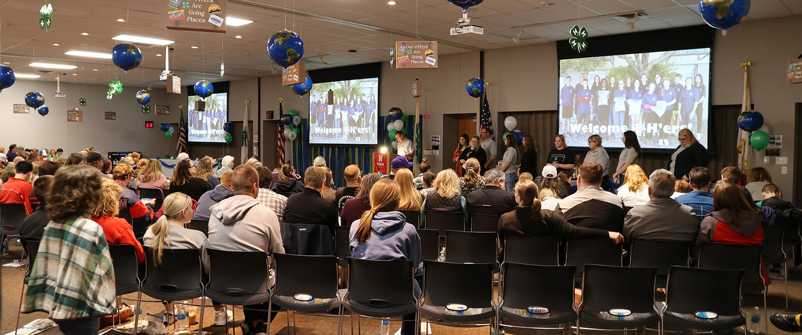 Room full of people sitting in chairs looking at three screens in front of the room, with globe and suitcase decorations hanging from the ceiling