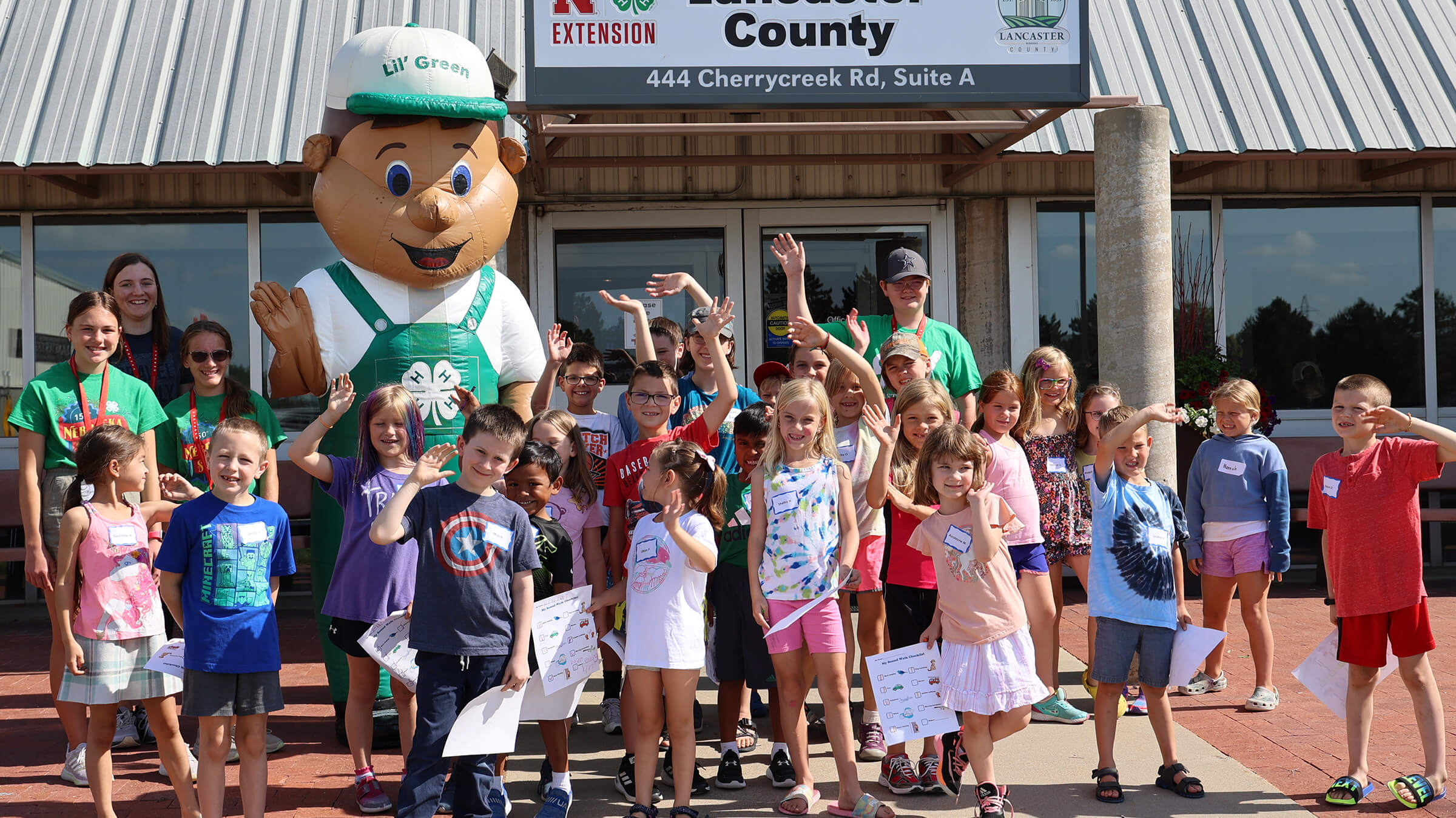 Lil Green inflatable mascot with group of youth in front of Extension building, all waving