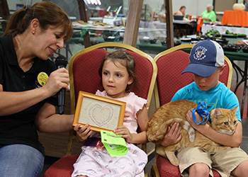 an adult talking to a girl about her drawing, and a boy is holding a cat next to them