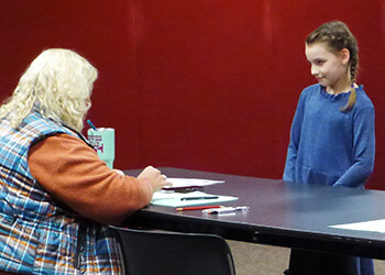 a young girl standing talking to an adult who is sitting at a table