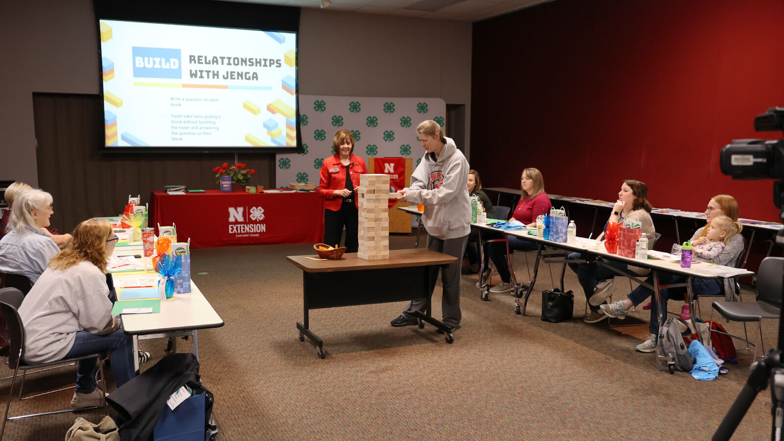 Several adult women at tables in a U shape watching a person in the center at a Jenga game. Screen in background says "Building Relationships With Jenga"