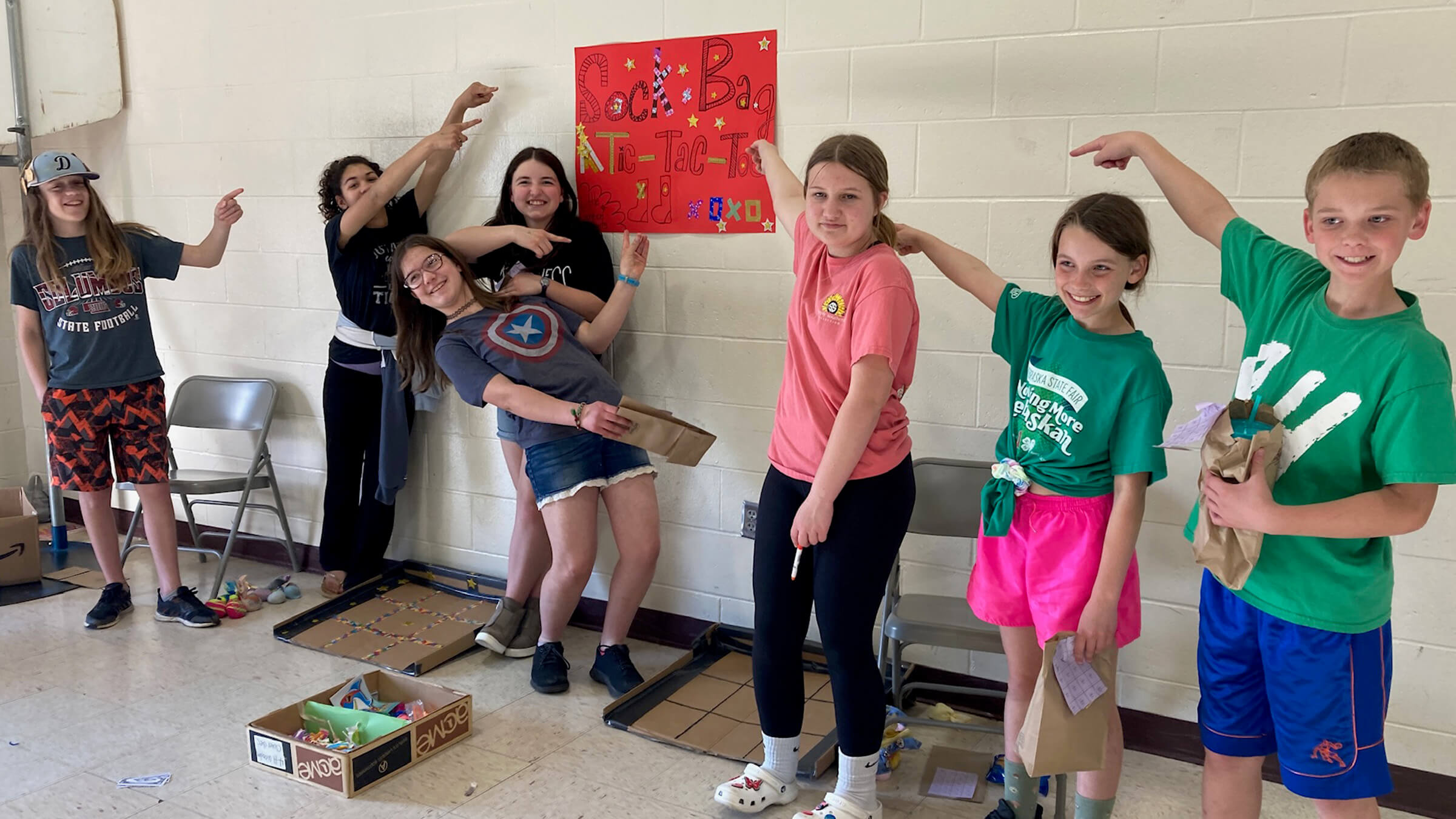 several boys and girls standing by cardboard tic tock toe boxes, pointing to a sign that says Sock Bag Tic Tac Toe