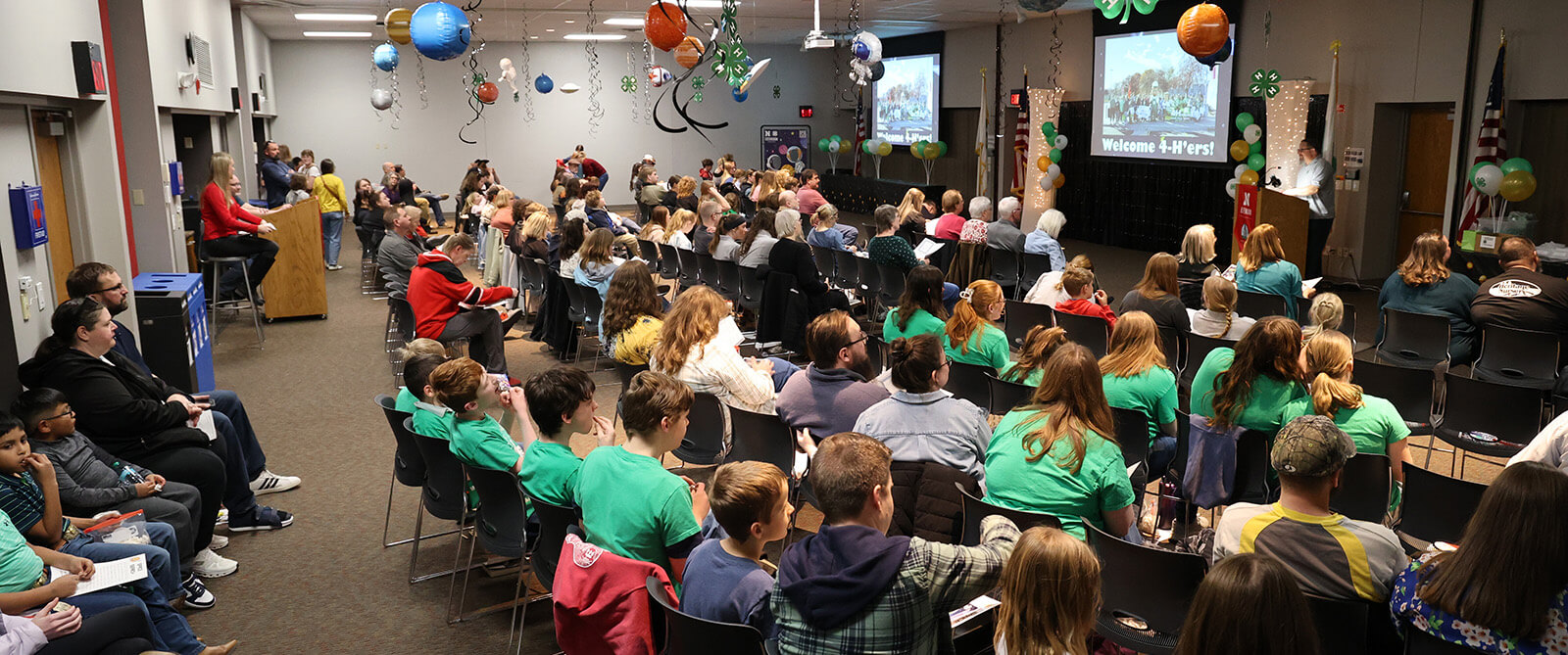 Festive decorated room full of adults and youth sitting in chairs looking at screens that say Welcome 4-H'ers