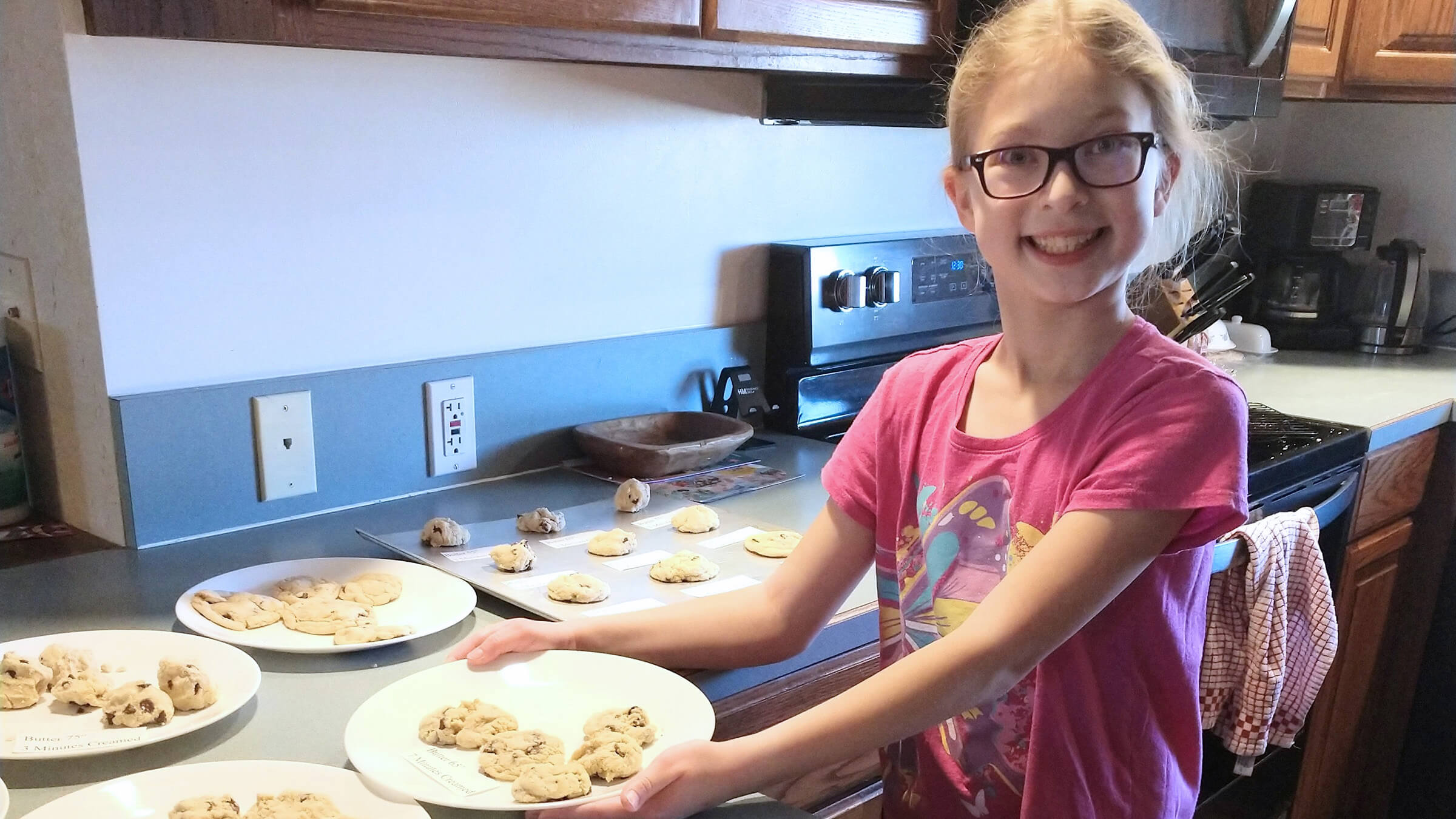 a girl in a kitchen with multiple batches of cookies which have labels