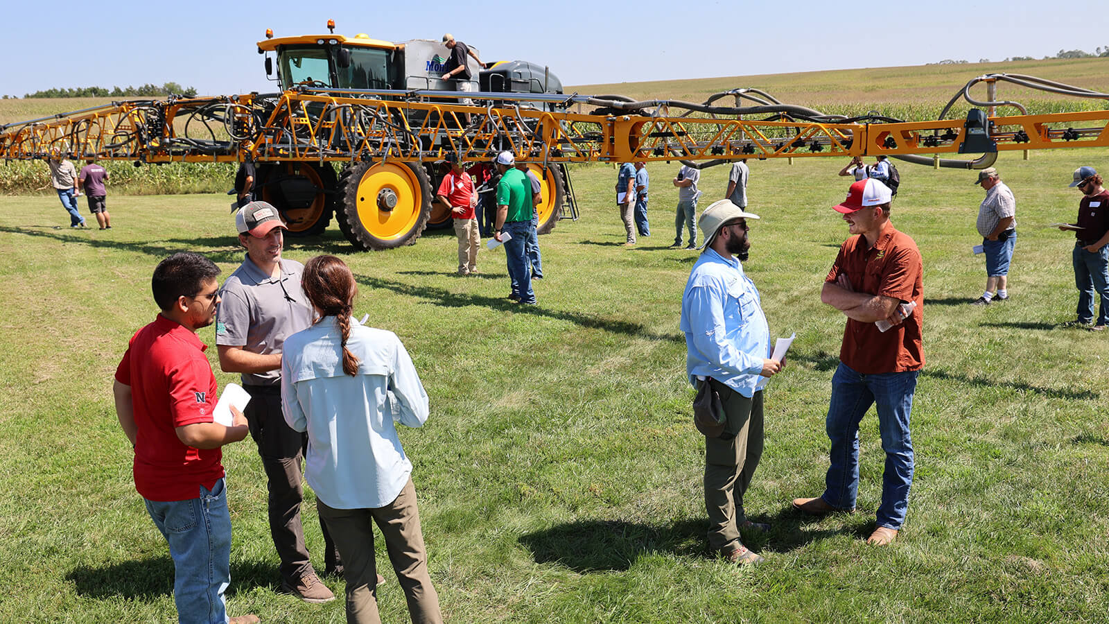 Several farmers talking in pairs or small groups with a large farm highboy machine in the background