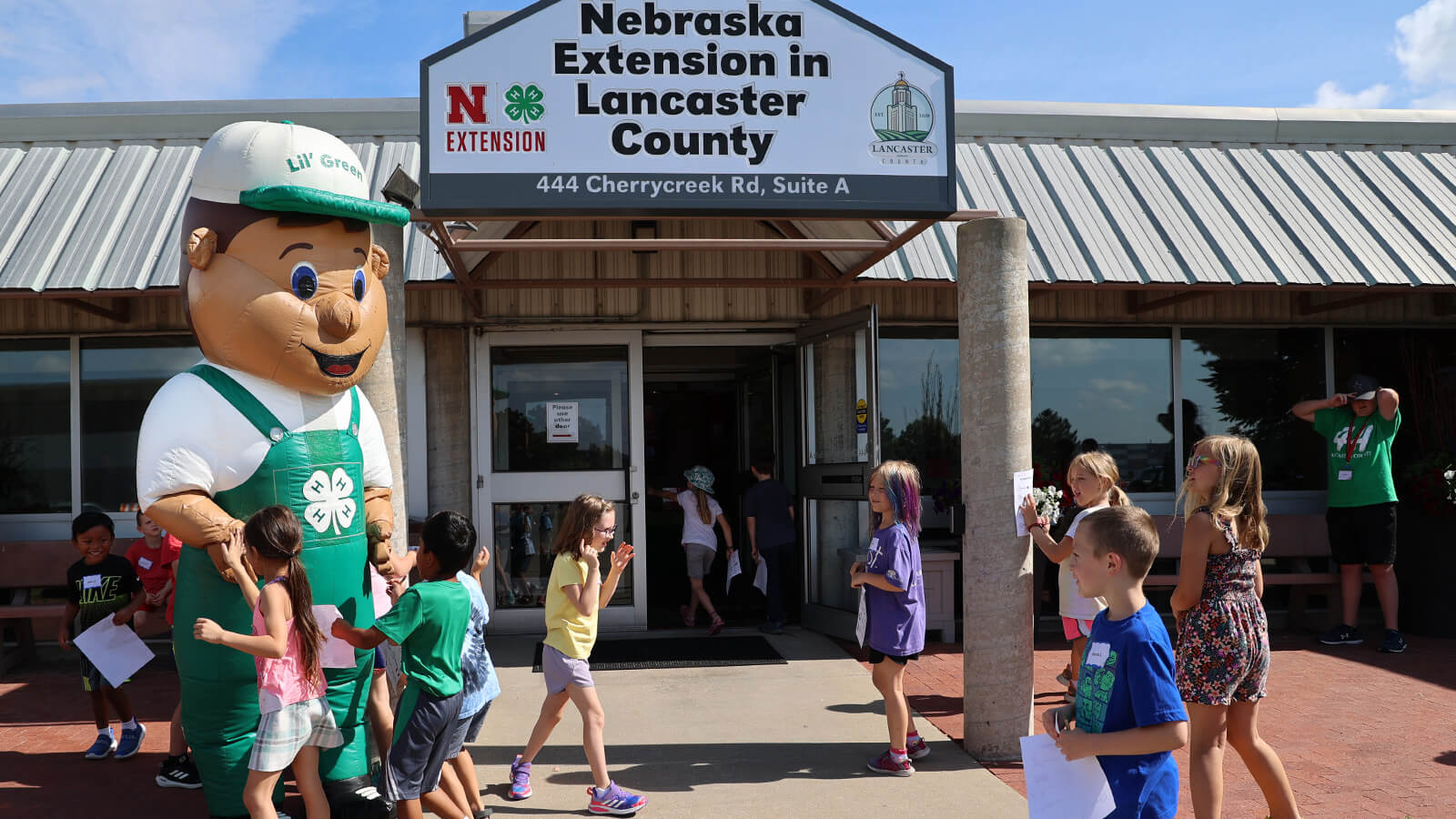 Lil Green blow up mascot giving high fives to young children in front of building which has a sign "Nebraska Extension in Lancaster County