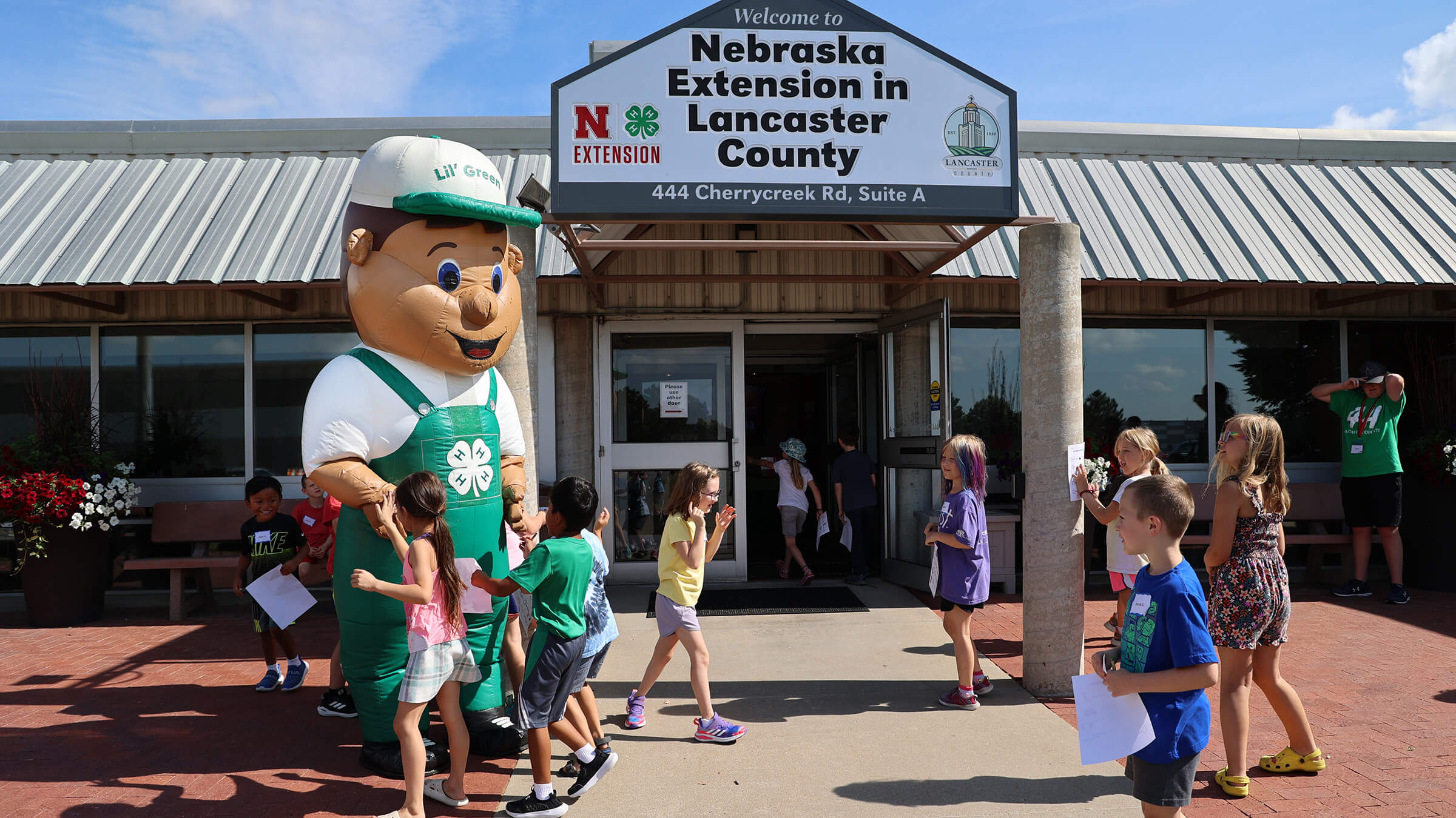 group of youth gathering around a blow-up mascot of Lil' Green in front of the Extension building