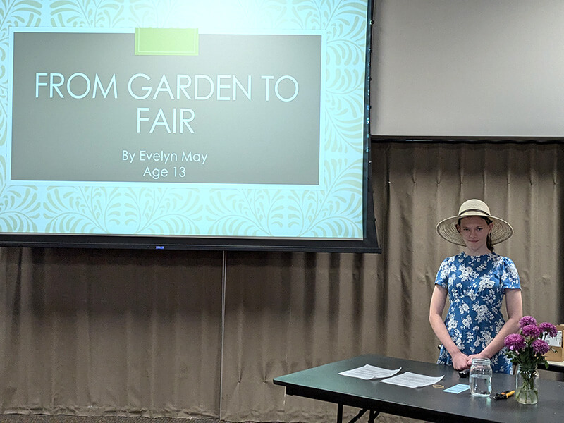 A teen girl in a floral pattern dress and garden hat standing by a projector screen that says From Garden to Fair