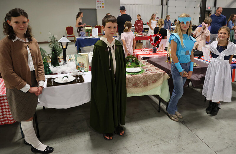 Three rows of youth in various costumes each standing next to a card table with table cloth, place setting and handmade centerpieces