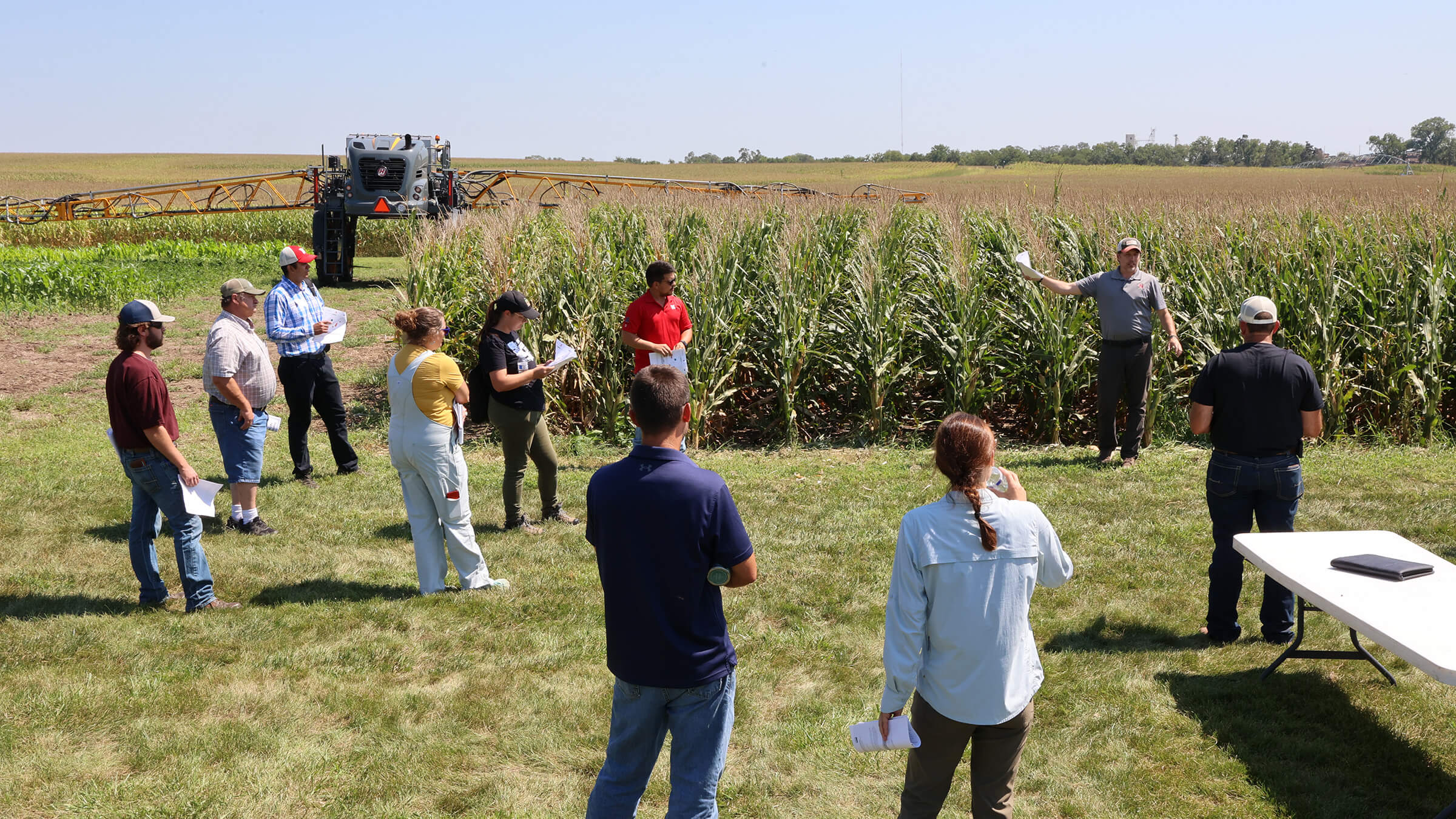 several people listening to an educator in front of a corn field with a large farm equipment in the background