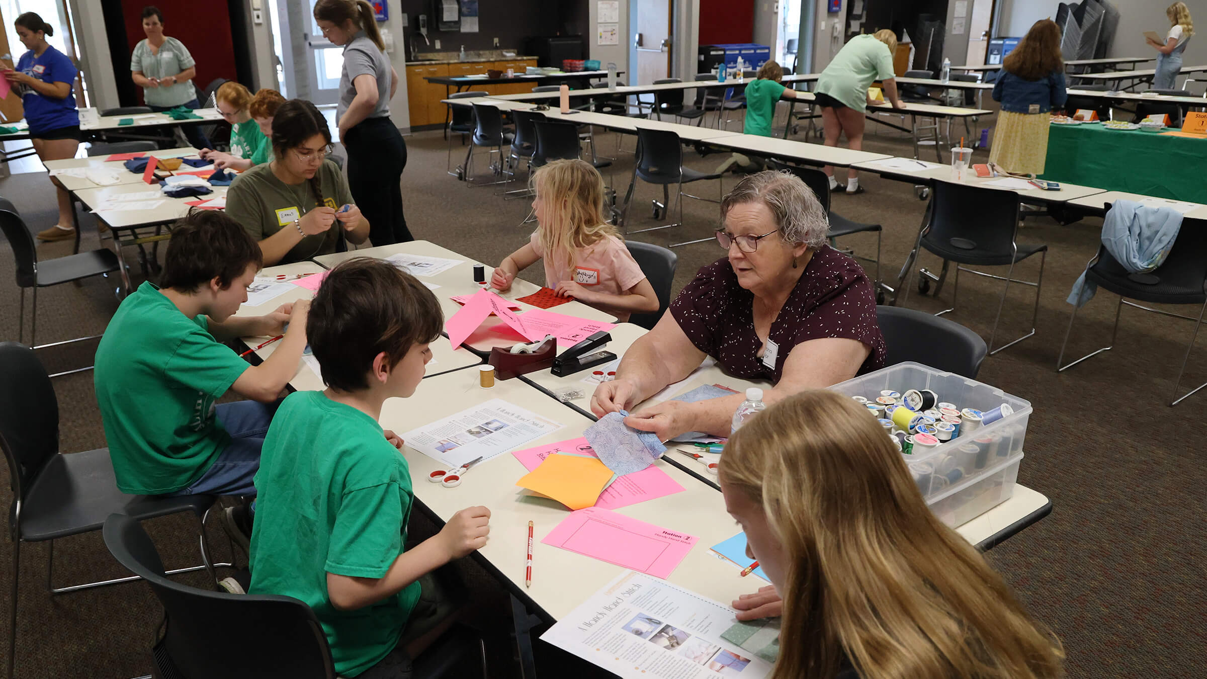 several youth around a table practicing a simple sewing stitch, led by an adult