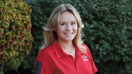 Olivia Lentfer in a red Nebraska Extension polo shirt standing outdoors with greenery in the background.