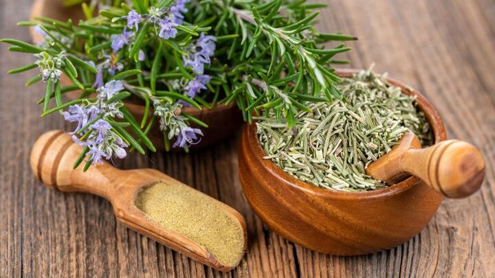 Fresh rosemary stems with small purple flowers next to a wooden bowl full of dried rosemary with a wooden scoop. An additional wooden scoop lays next to the fresh rosemary and wooden bowl. 