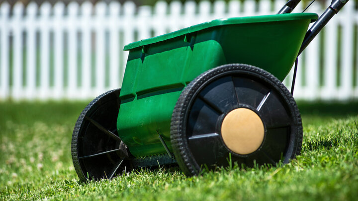 Drop seeder on green lawn near a white picket fence.