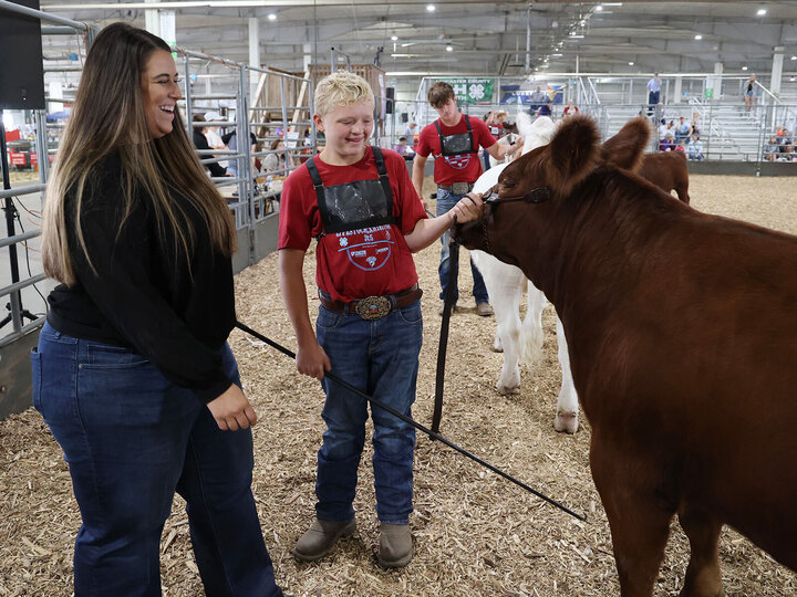 A young person holding a brown beef cow on a leash in a livestock arena, standing beside a smiling adult.
