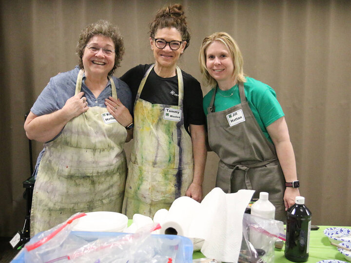 Three women smiling and wearing aprons behind a table with paint and paper towels