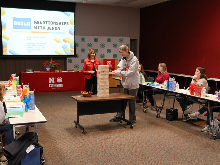 Several adult women at tables in a U shape watching a person in the center at a Jenga game. Screen in background says "Building Relationships With Jenga"