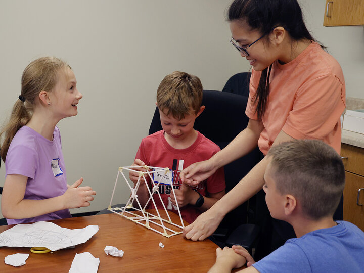A woman helping 3 youth building a bridge out of straws and tape