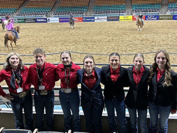 seven youth standing in front of an arena with riders on horses