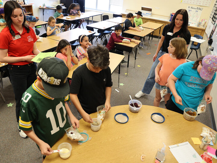 Two women looking over a row of students putting trail mix ingredients into bags