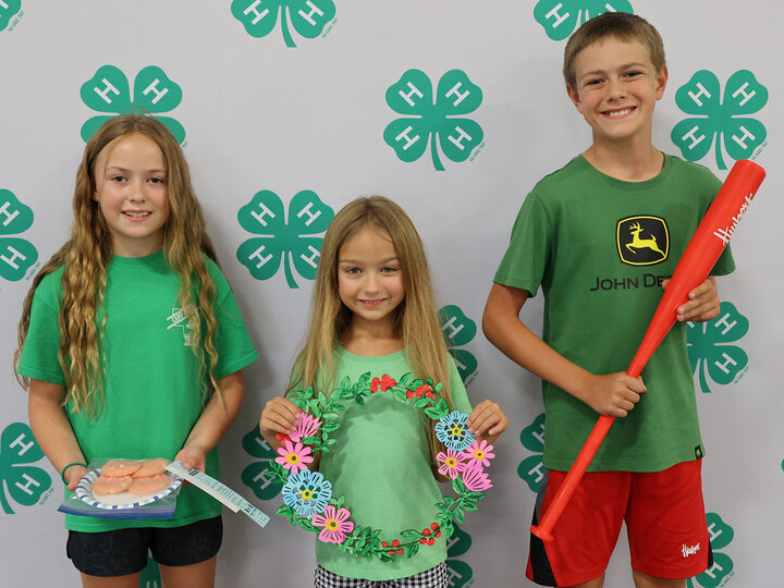 Two girls and a boy holding exhibits for the fair: cookies, a wall hanging, and a 3D printed baseball bat