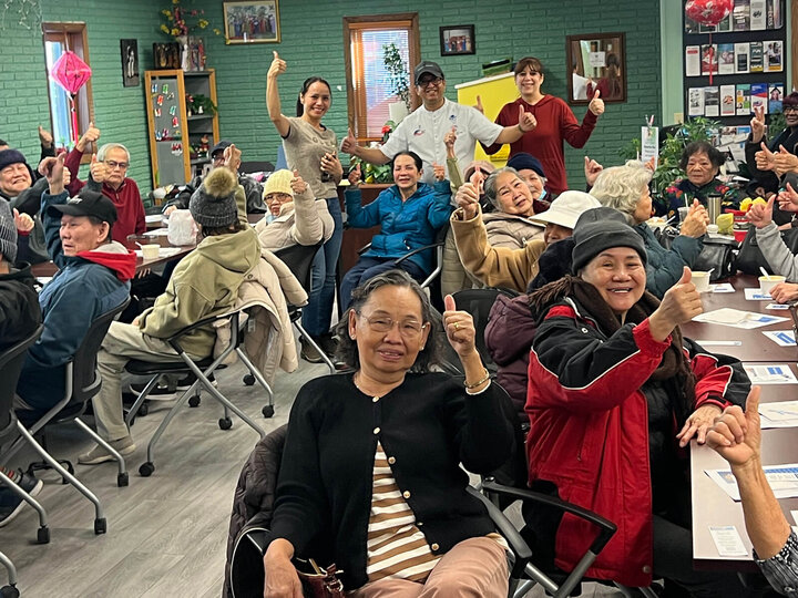 A room full of senior citizens sitting at tables doing thumbs up