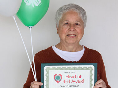 A smiling woman holding 2 balloons with 4-H logos and a certificate that says "Heart of 4-H Award"
