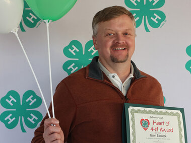 A man smiling holding 2 balloons and a certificate which reads "Heart of 4-H Award: Jason Babcock"