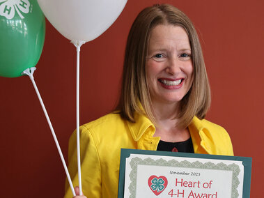 Woman holding a certificate which says "Heart of 4-H  Award" and two balloons with 4-H logo