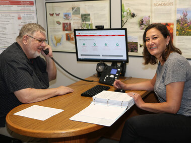 Two people sitting at a desk with a computer. Man is answering phone and woman has a 3-ring binder.