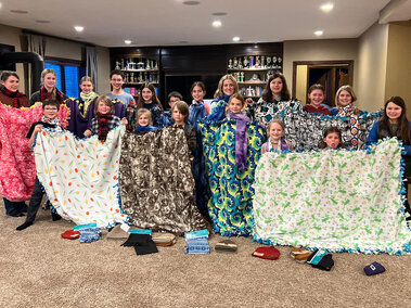 Two lines of youth holding large tie blankets up to display. Hats and scarfs are on the floor in front of them