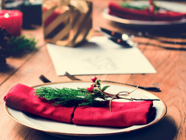 two plates with cloth napkins and tiny evergreen tree branches  on a table with candles and other greenery
