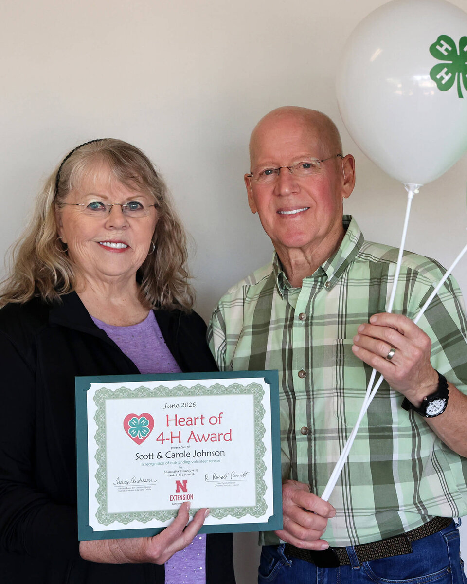 Man and woman holding a white balloon with 4-H clover emblem and an award certificate that says Heart of 4-H Award: Scott and Carole Johnson